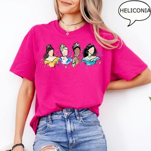 May include: A magenta t-shirt featuring a watercolor illustration of four women in gowns. The design includes yellow, blue, and teal accents. The word "HELICONIA" is in a speech bubble in the upper right corner.
