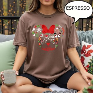 K&ouml;nnte beinhalten: Espressofarbenes T-Shirt mit festlichem Weihnachtsdesign. Das Design umfasst eine rote Schleife, Schneeflocken und die Worte "Joy to the World" und "Merry Christmas". Das Shirt hat eine bequeme Passform.
