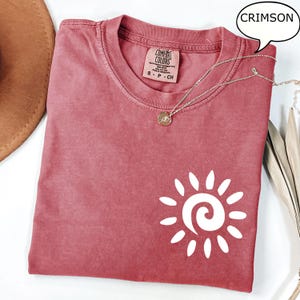 May include: A crimson colored t-shirt with a white sun graphic. The t-shirt has a distressed look and is made of a soft, comfortable fabric. The text "COMFORT COLORS" is visible on the tag.