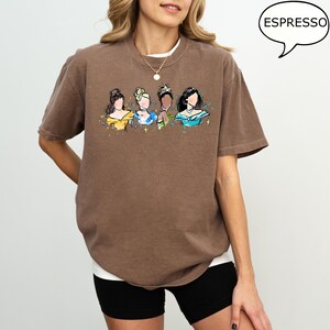 May include: Brown t-shirt with a watercolor illustration of four princesses. The princesses are depicted with yellow, blue, and green accents. The word "ESPRESSO" is in a speech bubble in the upper right corner.