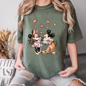 May include: A green t-shirt with a graphic of Mickey Mouse and Minnie Mouse holding hands with hearts floating around them.