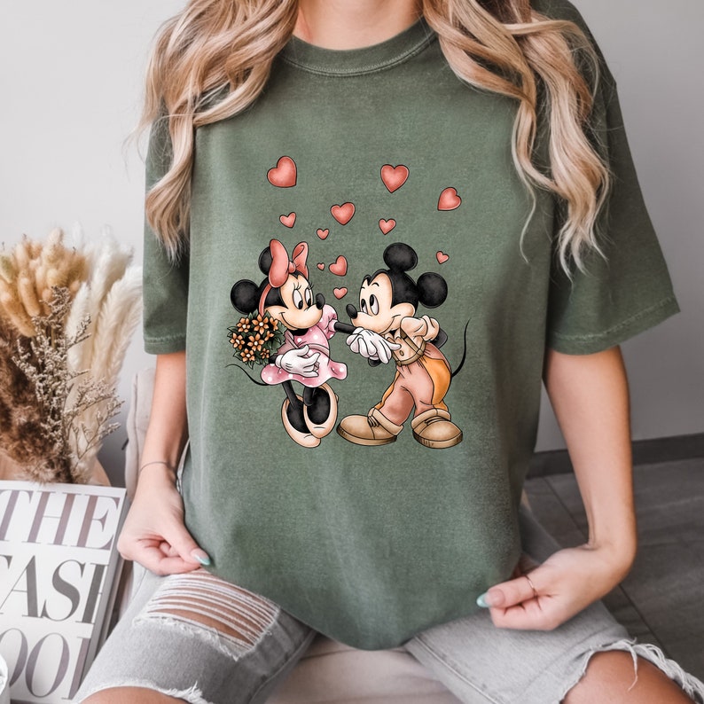 May include: A green t-shirt with a graphic of Mickey Mouse and Minnie Mouse holding hands with hearts floating around them.
