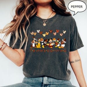 May include: Dark gray t-shirt featuring a Thanksgiving graphic with Disney characters. The design includes Mickey Mouse, Donald Duck, and other characters, along with the word "THANKSGIVING" in orange letters. The shirt has a relaxed fit.
