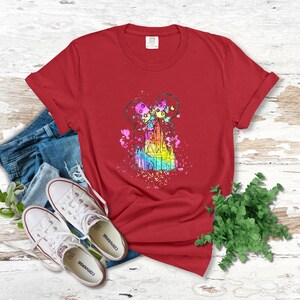 Puede incluir: Camiseta roja con una ilustraci&oacute;n de acuarela de un castillo con un esquema de colores arco&iacute;ris y orejas de Mickey Mouse. El castillo est&aacute; rodeado de salpicaduras de colores.