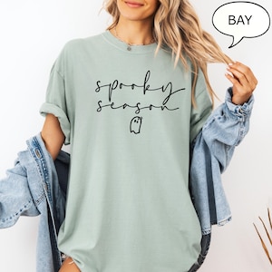 May include: A pale green t-shirt featuring the phrase "spooky season" in black cursive, and a small ghost graphic. The top has a crew neck and short sleeves. A denim jacket is worn over the shirt.