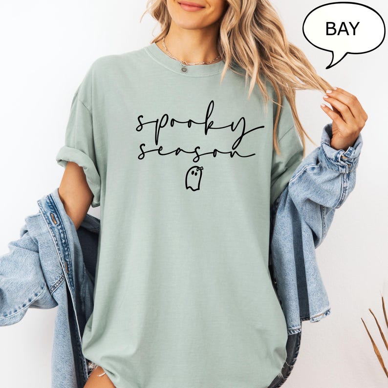 May include: A pale green t-shirt featuring the phrase "spooky season" in black cursive, and a small ghost graphic. The top has a crew neck and short sleeves. A denim jacket is worn over the shirt.
