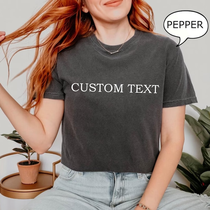 May include: A dark gray cropped t-shirt with the text "CUSTOM TEXT" printed in white on the front. The t-shirt is being worn by a person with red hair.
