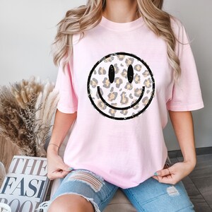 May include: A pink t-shirt with a distressed black and white smiley face graphic. The smiley face is filled with a leopard print pattern in shades of brown and yellow.