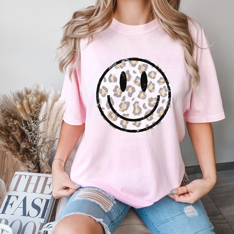 May include: A pink t-shirt with a distressed black and white smiley face graphic. The smiley face is filled with a leopard print pattern in shades of brown and yellow.