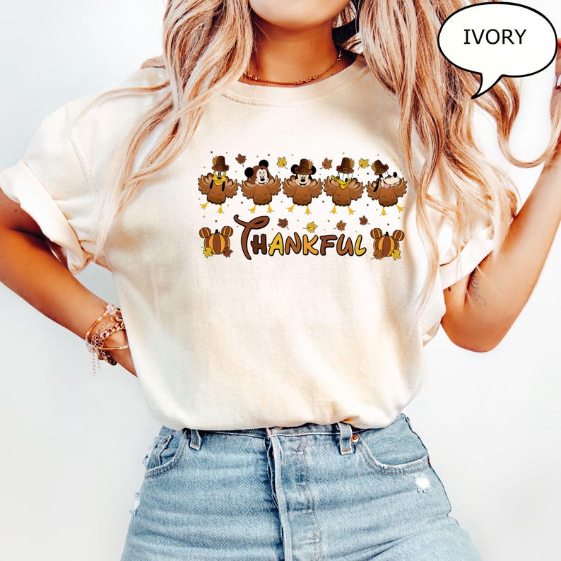 May include: Ivory-colored t-shirt featuring a Thanksgiving-themed design. The design includes cartoon turkeys wearing hats, with the word "Thankful" in a decorative font. The shirt is made of a soft material and is suitable for casual wear.
