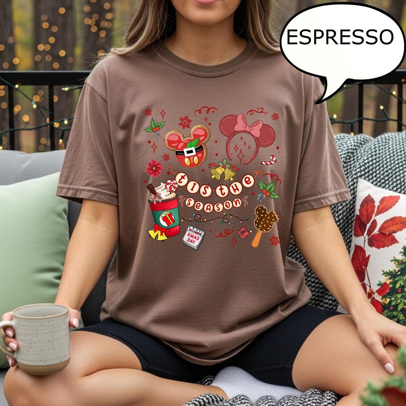 May include: A brown t-shirt with the text "'Tis the Season" and festive graphics. The design includes a gingerbread Mickey Mouse, a Minnie Mouse headband, and a Christmas-themed drink. The shirt is a relaxed fit and the color is described as Espresso.