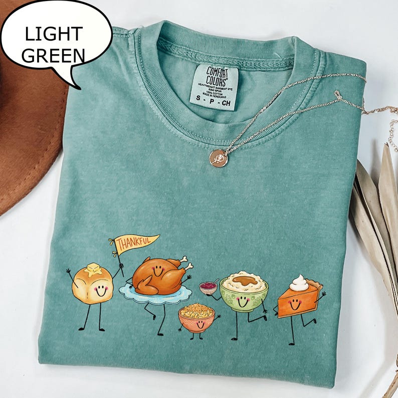 Pu&ograve; includere: Una t-shirt a maniche corte verde chiaro con una grafica di cibi del Ringraziamento antropomorfi, tra cui un panino che tiene una bandierina con la scritta THANKFUL, un tacchino arrosto, una ciotola di ripieno, pur&egrave; di patate e una fetta di torta.