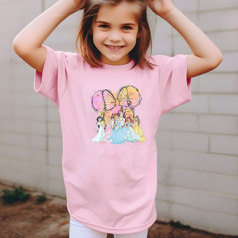 Puede incluir: Una camiseta rosa claro con un gr&aacute;fico colorido de un lazo con orejas de rat&oacute;n y varias princesas frente a un castillo. El gr&aacute;fico est&aacute; centrado en la parte delantera de la camiseta.
