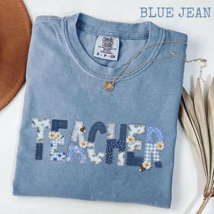May include: A blue jean coloured t-shirt with a crew neckline and a Comfort Colors label. The word TEACHER is appliqued across the chest in various blue and white patterned fabrics, including gingham and floral prints, accented with small daisies.