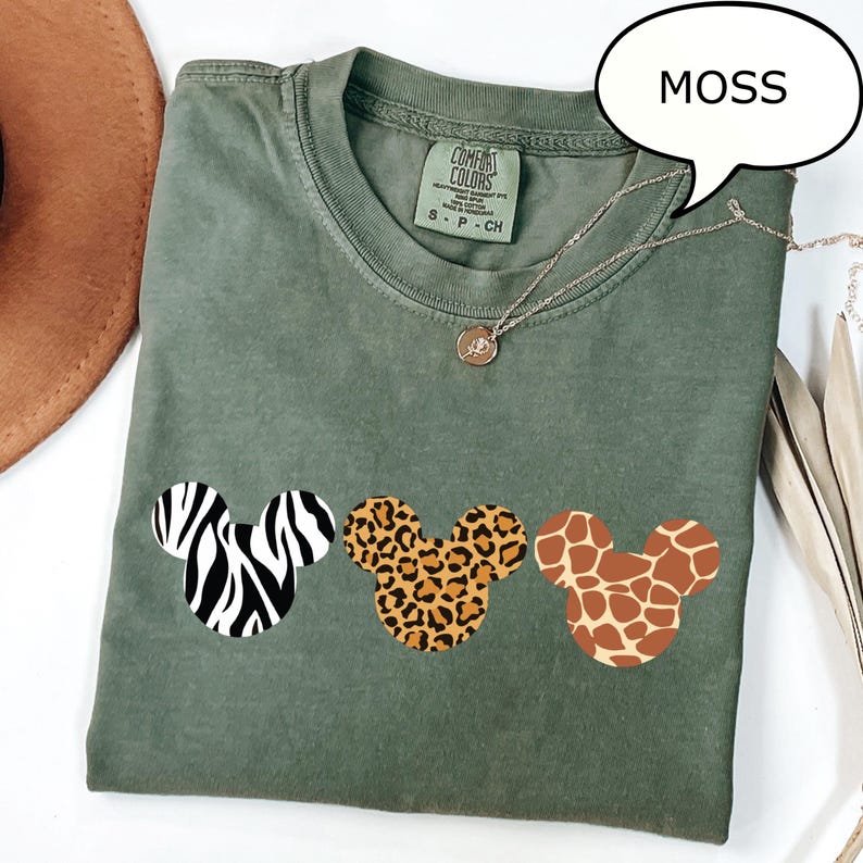 Pu&ograve; includere: T-shirt verde muschio con tre disegni a testa di Topolino con motivi zebrati, leopardati e giraffa. La maglietta ha girocollo e maniche corte. Una collana d'argento con un ciondolo &egrave; drappeggiata sulla maglietta.