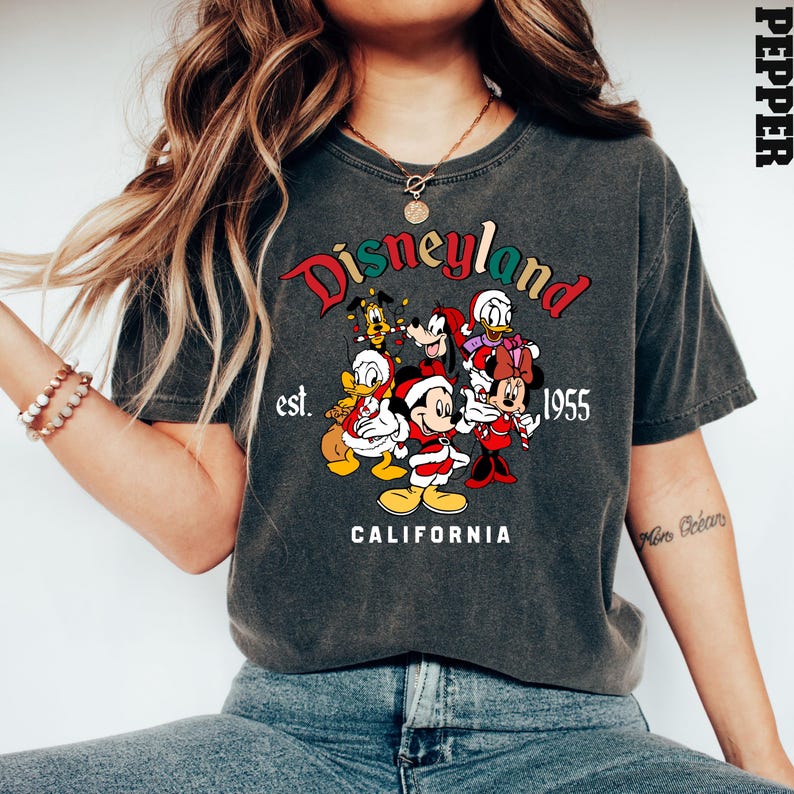 May include: A gray t-shirt with a vintage-style graphic of Mickey Mouse and friends in front of a Disneyland sign. The text on the shirt reads "Disneyland est. 1955 California".