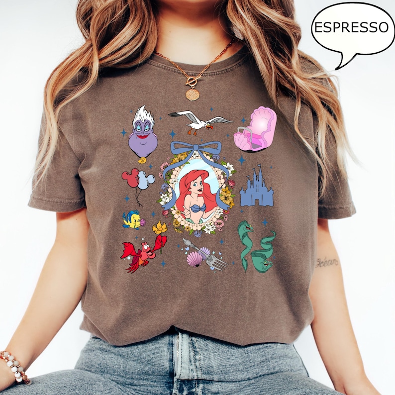 May include: A brown t-shirt with a graphic print of various characters from the Disney movie The Little Mermaid. The characters include Ariel, Ursula, Sebastian, Flounder, and the sea witch. The graphic also includes a castle, a seashell, and a trident.