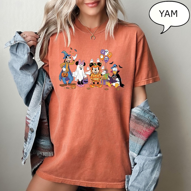 May include: Orange t-shirt featuring a Halloween-themed graphic of Disney characters, including Mickey Mouse, Minnie Mouse, Donald Duck, and Goofy, dressed in costumes. The graphic includes pumpkins, ghosts, and bats. The word "YAM" is in a speech bubble.