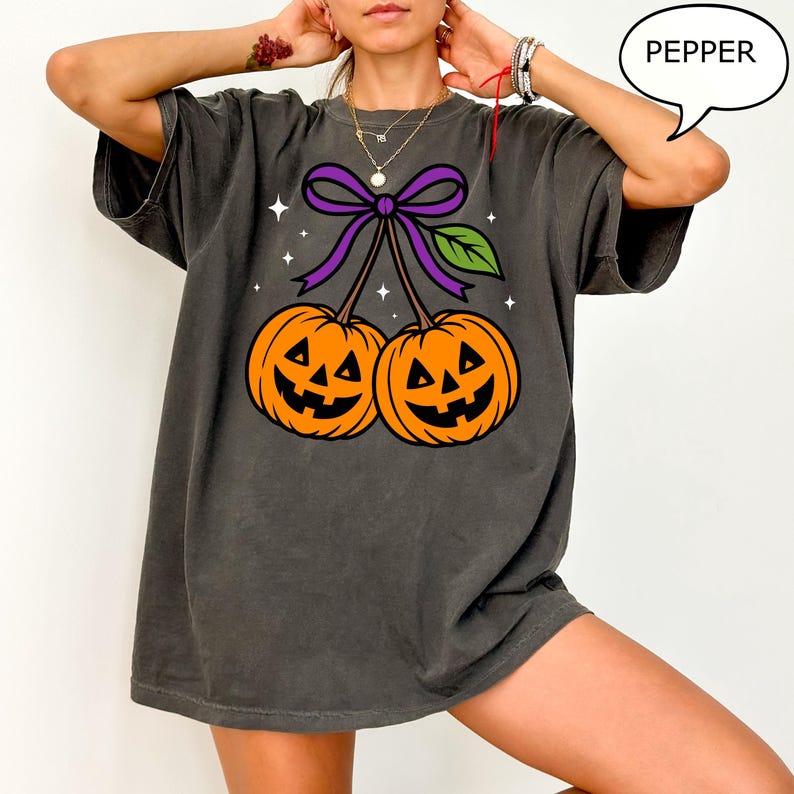 May include: Dark grey t-shirt featuring a Halloween-themed graphic. The design includes two orange jack-o'-lanterns connected by a purple bow and green leaf. The word "PEPPER" is in a speech bubble. The shirt is a casual, oversized fit.