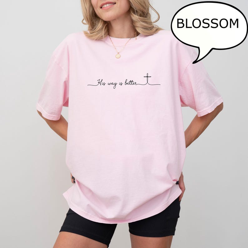 May include: A light pink t-shirt with the text "His way is better" and a cross design. The word "BLOSSOM" is in a speech bubble in the upper right corner. The shirt is worn by a person.