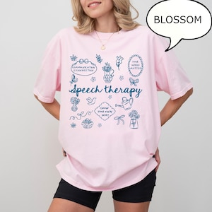 May include: A light pink t-shirt with the word "Speech therapy" in a script font, surrounded by blue illustrations of flowers, a watering can, and other related graphics. The shirt also has the text "Communication is connection" and "Your words matter".