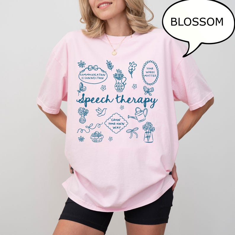 May include: A light pink t-shirt with the word "Speech therapy" in a script font, surrounded by blue illustrations of flowers, a watering can, and other related graphics. The shirt also has the text "Communication is connection" and "Your words matter".