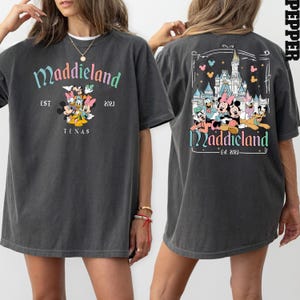 K&ouml;nnte beinhalten: Ein graues Oversize-T-Shirt mit einem grafischen Druck von Mickey Mouse und seinen Freunden vor einem Schloss. Der Text "Maddieland Est. 2023 Texas" ist auf der Vorderseite des Shirts gedruckt. Die R&uuml;ckseite des Shirts hat einen &auml;hnlichen grafischen Druck mit dem Text "Maddieland Est. 2023".