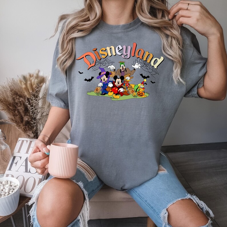 Puede incluir: Una camiseta gris con un gr&aacute;fico de Mickey Mouse y sus amigos vestidos con disfraces de Halloween. El texto "Disneyland" est&aacute; impreso en colores arco&iacute;ris sobre el gr&aacute;fico.