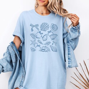 May include: A light blue t-shirt with a pattern of blue seashells and starfish. The design is centered on the shirt.