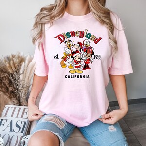 May include: Pink t-shirt with a graphic print of Mickey Mouse and friends in front of a Disneyland logo that says "Disneyland est. 1955 California".