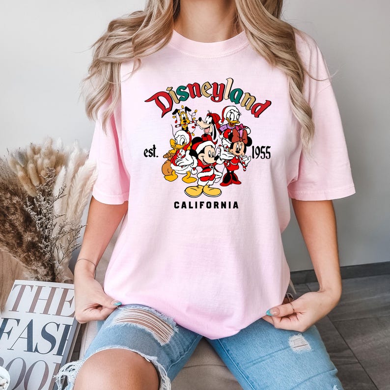 May include: Pink t-shirt with a graphic print of Mickey Mouse and friends in front of a Disneyland logo that says "Disneyland est. 1955 California".