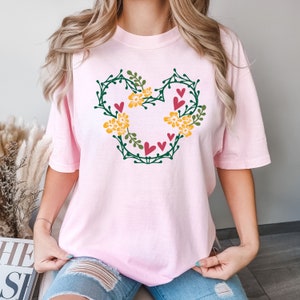 K&ouml;nnte beinhalten: Ein pinkfarbenes T-Shirt mit einem floralen Mickey Mouse-Design. Das Design enth&auml;lt gr&uuml;ne Bl&auml;tter, gelbe Blumen und rosa Herzen.