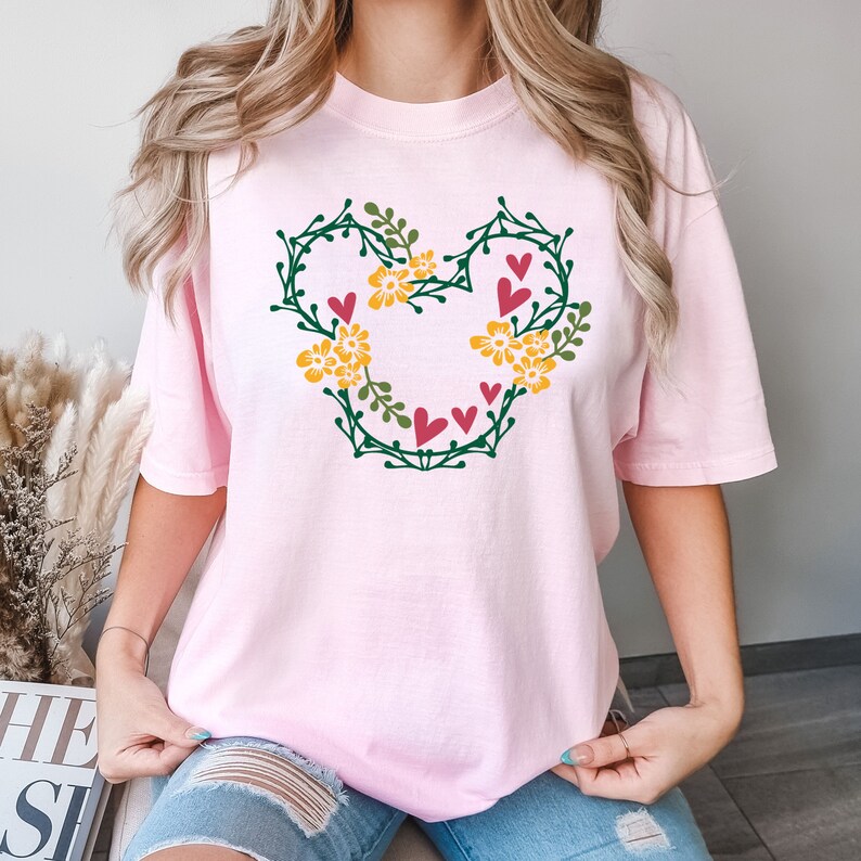 K&ouml;nnte beinhalten: Ein pinkfarbenes T-Shirt mit einem floralen Mickey Mouse-Design. Das Design enth&auml;lt gr&uuml;ne Bl&auml;tter, gelbe Blumen und rosa Herzen.
