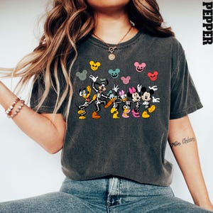 May include: A dark gray t-shirt with a graphic print of Mickey Mouse, Minnie Mouse, Donald Duck, and Goofy dressed as skeletons holding colorful balloons. The text "Pepper" is visible in the upper right corner of the image.