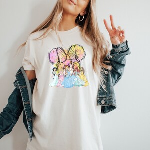 Puede incluir: Camiseta color crema con un gr&aacute;fico colorido de un castillo y varias princesas. El dise&ntilde;o incluye un gran lazo con acentos amarillos y rosas. La camiseta la lleva una persona.