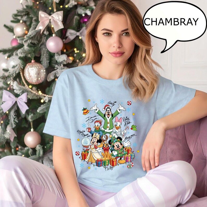 Puede incluir: Camiseta azul claro con un dise&ntilde;o navide&ntilde;o de personajes de Disney. El gr&aacute;fico incluye a Mickey y Minnie Mouse, Donald Duck, Daisy Duck, Goofy y Pluto, rodeados de regalos y decoraciones. La palabra "CHAMBRAY" est&aacute; en un bocadillo.