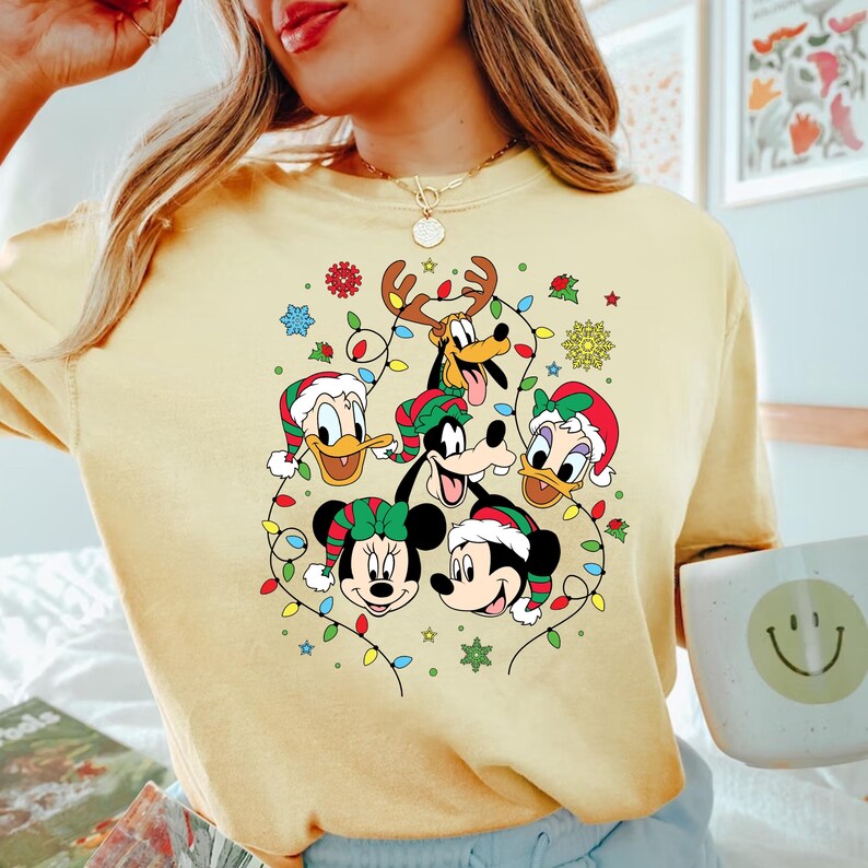 May include: A yellow t-shirt with a Christmas-themed design featuring Mickey Mouse, Minnie Mouse, Donald Duck, Goofy, and Pluto. The characters are surrounded by colorful Christmas lights and snowflakes.