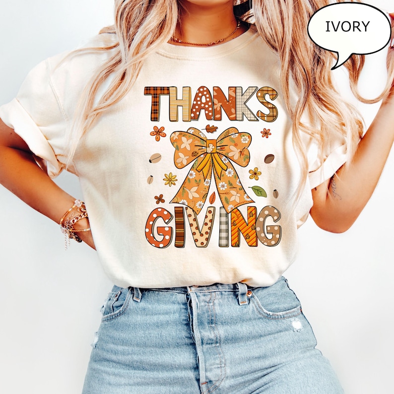 May include: Ivory-colored t-shirt with a Thanksgiving theme. The shirt features the words "THANKS GIVING" in a patchwork design, a large bow, and floral and leaf accents. The shirt is a casual, festive garment.