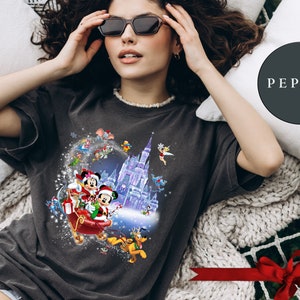 Puede incluir: Una camiseta gris oscuro con un dise&ntilde;o navide&ntilde;o que presenta a Mickey Mouse y Minnie Mouse montando en un trineo con un castillo morado en el fondo. El dise&ntilde;o tambi&eacute;n incluye otros personajes de Disney como Pluto, Donald Duck y Goofy.