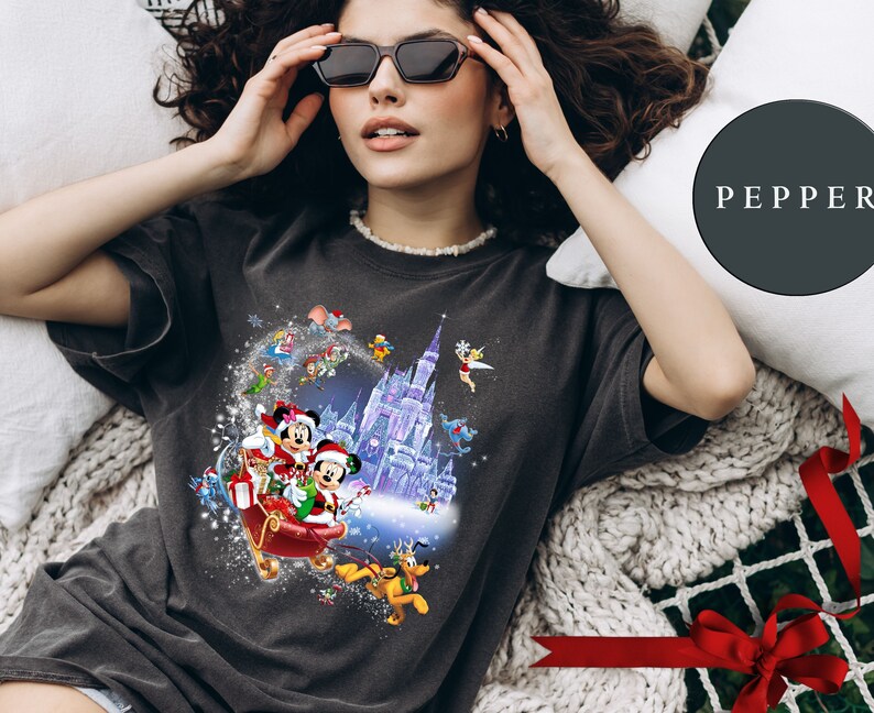 Puede incluir: Una camiseta gris oscuro con un dise&ntilde;o navide&ntilde;o que presenta a Mickey Mouse y Minnie Mouse montando en un trineo con un castillo morado en el fondo. El dise&ntilde;o tambi&eacute;n incluye otros personajes de Disney como Pluto, Donald Duck y Goofy.