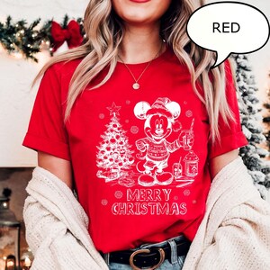 Puede incluir: Camiseta roja con un gr&aacute;fico blanco de Mickey Mouse con gorro de Pap&aacute; Noel, sosteniendo una bebida y junto a un &aacute;rbol de Navidad. El texto "MERRY CHRISTMAS" est&aacute; debajo. La palabra "RED" est&aacute; en una burbuja.