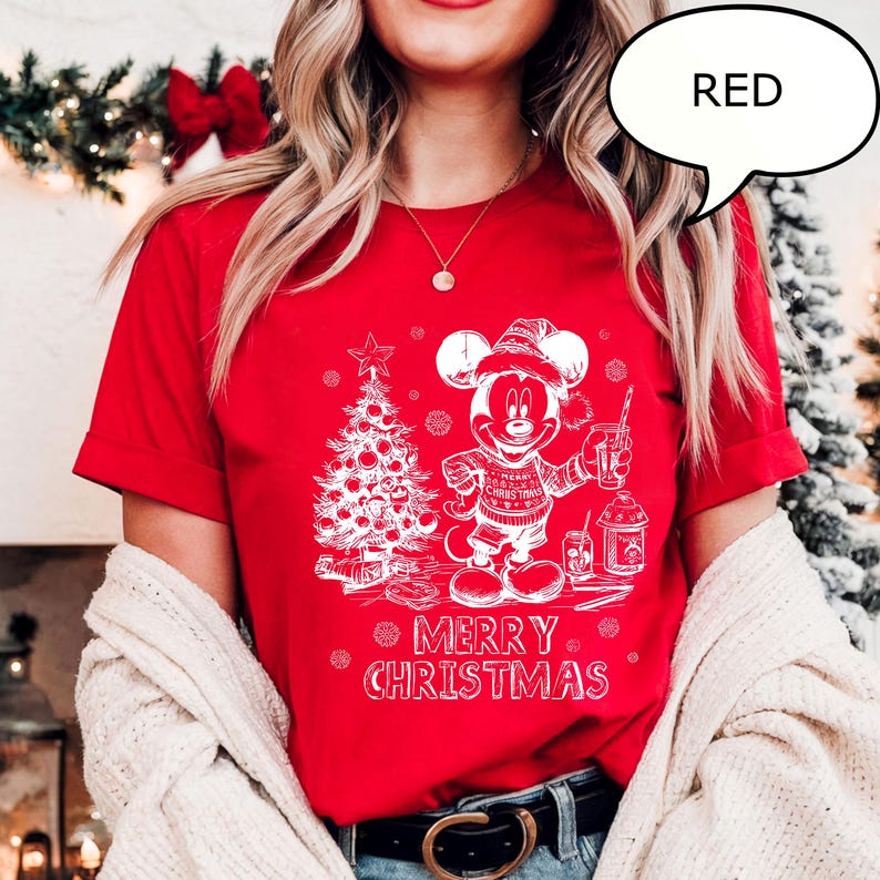Puede incluir: Camiseta roja con un gr&aacute;fico blanco de Mickey Mouse con gorro de Pap&aacute; Noel, sosteniendo una bebida y junto a un &aacute;rbol de Navidad. El texto "MERRY CHRISTMAS" est&aacute; debajo. La palabra "RED" est&aacute; en una burbuja.