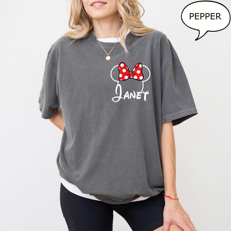 May include: Gray t-shirt with a white collar featuring a Minnie Mouse design. The design includes a red bow with white polka dots, mouse ears, and the name "Janet" in white script. A speech bubble with the word "Pepper" is in the upper right corner.