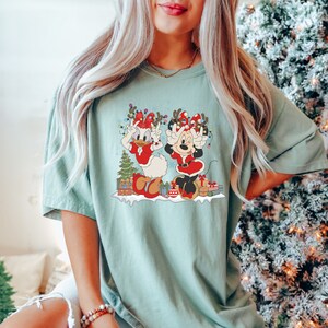 Puede incluir: Una camiseta azul turquesa con un gr&aacute;fico navide&ntilde;o que presenta a Minnie Mouse y Daisy Duck con gorros de Pap&aacute; Noel y tap&aacute;ndose los ojos con las manos. El gr&aacute;fico tambi&eacute;n incluye un &aacute;rbol de Navidad, regalos y luces.