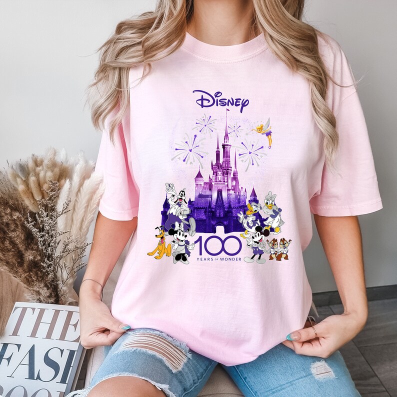 May include: Pink t-shirt with a purple and white graphic of Cinderella's castle, fireworks, and Disney characters. The text "Disney 100 Years of Wonder" is printed on the shirt.