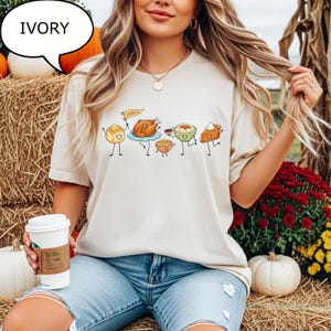 Pu&ograve; includere: Una t-shirt color avorio con una stampa grafica di cibi del Ringraziamento antropomorfi, tra cui un tacchino arrosto che tiene una bandierina gialla con la scritta THANKFUL, un panino, una ciotola di pur&egrave; di patate, ripieno e una fetta di torta di zucca.