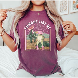 May include: A purple t-shirt with a graphic of a frog wearing a pink cowboy hat and holding a watermelon slice. The text "COWBOY LIKE ME" is printed above the frog.