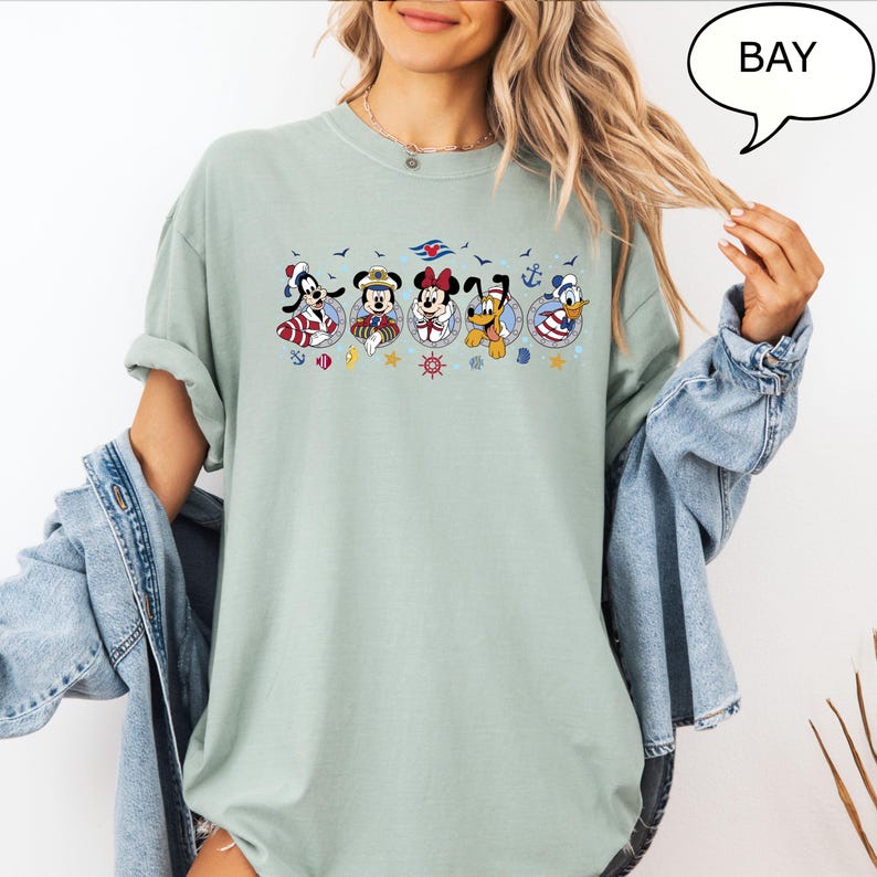 May include: A light green t-shirt featuring a nautical-themed graphic of Disney characters, including Mickey, Minnie, Donald, and Goofy. The graphic includes anchors, stars, and the text "BAY" in a speech bubble. The shirt has rolled-up sleeves.