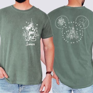 K&ouml;nnte beinhalten: Salbeigr&uuml;nes T-Shirt mit wei&szlig;er Grafik von Mickey Mouse vor einem Schloss, darunter der Name "James". Auf der R&uuml;ckseite des Shirts steht der Text "The Happiest Place on Earth" in einem Mickey Mouse-Ohren-Design.