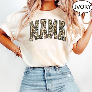 May include: A white t-shirt with the word "MAMA" printed in a camouflage pattern. The shirt is being worn by a person with long blonde hair and is available in the color ivory.